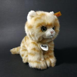 May include: A plush orange tabby cat with green eyes and a white muzzle. The stuffed animal has a round head and is sitting with its tail curled. A white tag with the word "Steiff" is attached to the cat's chest.