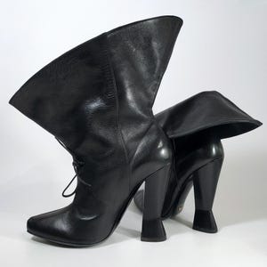 May include: A pair of black leather boots with a unique design. The boots have a high heel and a folded-over top. The boots have a lace-up closure and a pointed toe. The boots are set against a white background.