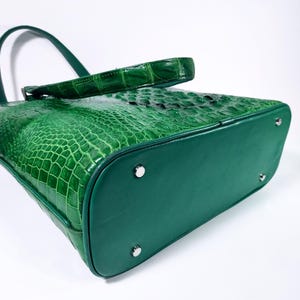 Pu&ograve; includere: Una borsa a mano verde con texture di coccodrillo, manico e base abbinati. La borsa presenta un design strutturato con piedini in metallo argentato. La pelle ha una finitura lucida e la borsa &egrave; su sfondo bianco.