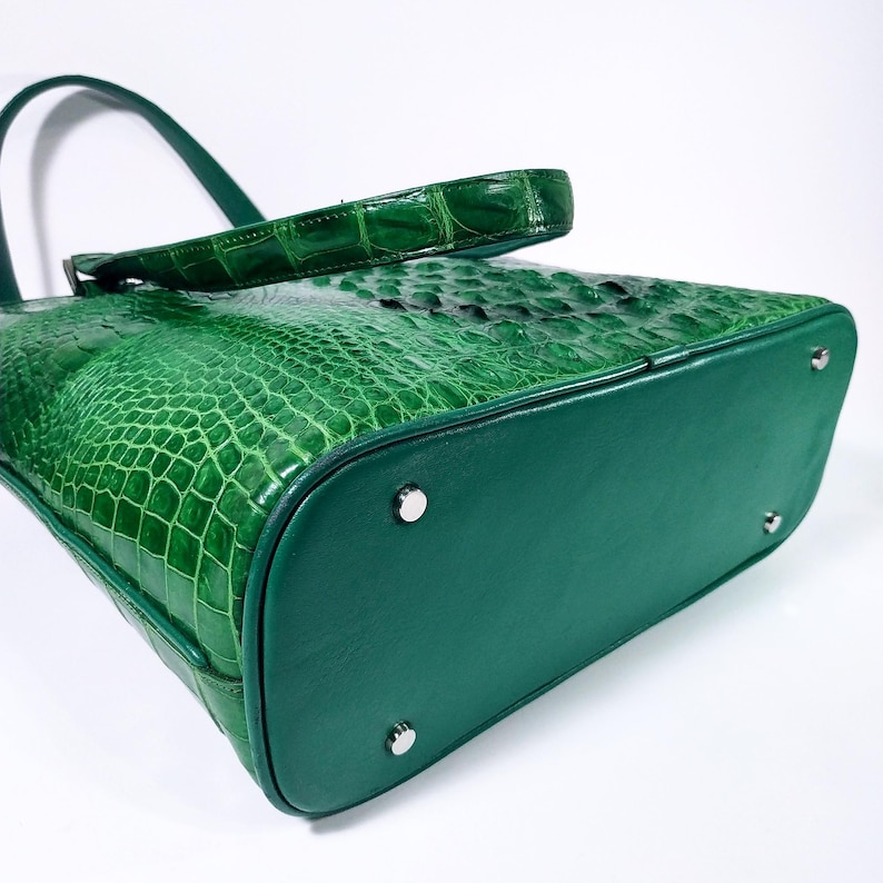 Pu&ograve; includere: Una borsa a mano verde con texture di coccodrillo, manico e base abbinati. La borsa presenta un design strutturato con piedini in metallo argentato. La pelle ha una finitura lucida e la borsa &egrave; su sfondo bianco.