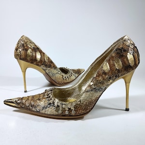 Può includere: Un paio di décolleté a punta con un motivo testurizzato in pelle di serpente nei toni dell'oro, marrone e crema. Le scarpe presentano un tacco a spillo color oro metallizzato, un puntale in metallo dorato e un bordo decorativo di cristalli.