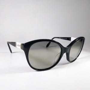 May include: Black sunglasses with a rounded frame and grey gradient lenses. The temples feature a white rectangular accent with the word Moschino printed in black. The frame is made of a glossy black material with a smooth finish and curved arms.