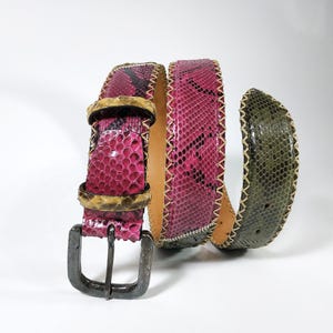 May include: A pink and green snakeskin belt with a dark metal buckle and gold-toned hardware. The belt has a stitched edge and a natural leather interior. The snakeskin pattern includes black accents.