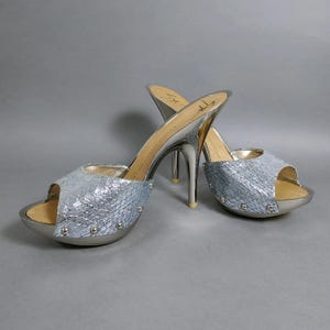 May include: A pair of silver metallic high-heeled mules featuring a textured snakeskin-patterned upper, an open-toe design, and a platform sole accented with small silver studs. The interior footbed displays the Giuseppe Zanotti brand signature.
