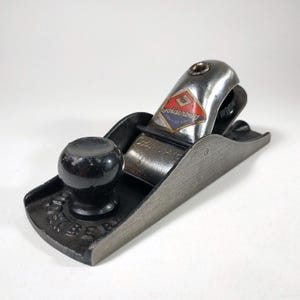 Może przedstawiać: Zabytkowy hebel ręczny z czarnym pokrętłem i srebrnym metalowym korpusem. Hebel ma nazwę "Goldenberg" i logo na górze. Podstawa hebla ma wygrawerowany napis "SWISS MADE".