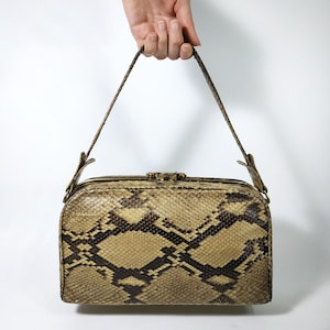 Könnte beinhalten: Eine rechteckige Handtasche mit Schlangenmuster in Beige-, Braun- und Schwarztönen. Die Tasche hat einen goldfarbenen Verschluss und einen Henkel. Die Tasche wird von einer Hand vor weißem Hintergrund gehalten.