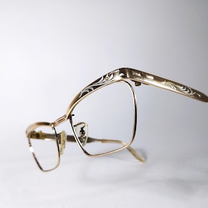 Puede incluir: Gafas doradas con un patrón floral decorativo en las patillas. Las monturas tienen un diseño clásico y vintage con forma de lente rectangular. Las gafas están sobre un fondo blanco.