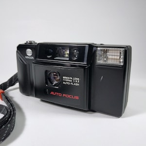 以下が含まれることがあります： 黒色のミノルタ35mmフィルムカメラ。内蔵フラッシュ付き。「MINOLTA LENS 1=35mm 1:3.5 AUTO FLASH」と「AUTO FOCUS」の文字が赤色で表示されています。黒とグレーのストラップが付いています。