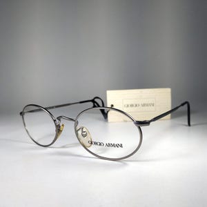May include: A pair of Giorgio Armani eyeglasses with oval frames and dark temples. The glasses have a silver-tone finish and clear lenses. The brand name is printed on the lens. A tag with the brand name is in the background.
