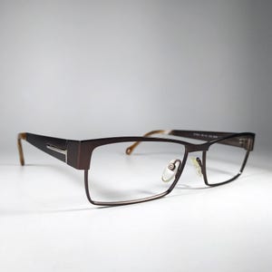 Könnte beinhalten: Braune rechteckige Brille mit dunkelbraunem Rahmen und hellbraunen Bügeln. Die Brille hat klare Gläser und einen silbernen Akzent an den Bügeln. Auf weißem Hintergrund.