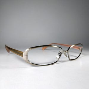 May include: Oval-shaped eyeglasses featuring a metallic silver frame with a brushed finish. The temples are a contrasting light brown wood-grain pattern, while the inner temple tips are a soft peach color. Includes adjustable nose pads.