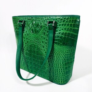 Pu&ograve; includere: Una borsa tote verde brillante con una texture di pelle di coccodrillo. La borsa presenta due lunghi manici verdi abbinati e finiture in metallo color argento. La borsa &egrave; rettangolare con una parte superiore leggermente arrotondata.