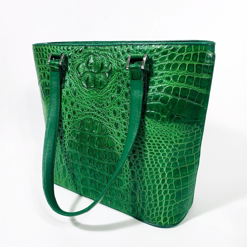 Pu&ograve; includere: Una borsa tote verde brillante con una texture di pelle di coccodrillo. La borsa presenta due lunghi manici verdi abbinati e finiture in metallo color argento. La borsa &egrave; rettangolare con una parte superiore leggermente arrotondata.