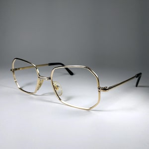 May include: Gold-rimmed eyeglasses with a geometric, octagonal shape. The frames are a shiny gold color, with black temples and nose pads. The lenses are clear. The glasses are set against a white background.