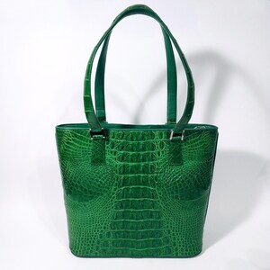 Pu&ograve; includere: Una borsa tote verde brillante con una texture a pelle di coccodrillo. La borsa ha due manici e una forma rettangolare. Il materiale &egrave; lucido e strutturato. I manici sono fissati con elementi metallici color argento.