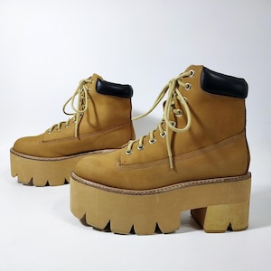 May include: A pair of tan platform boots with black padded collars and light-colored laces. The boots have a chunky, lugged sole and a stacked heel. The boots are made of a durable material and are designed for style and comfort.