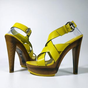 Peut inclure: Une paire de sandales à plateforme en cuir jaune vif avec des brides croisées, un haut talon en bois et une boucle de fermeture en métal argenté à la cheville. La semelle intérieure porte la marque Gucci et la pointure 37,5.