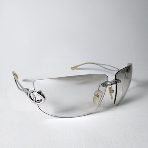 May include: Rimless sunglasses with a silver frame and tinted lenses. The sunglasses have a subtle, curved design with a small logo detail on the temples. The lenses are a gradient tint, transitioning from a darker shade at the top to a lighter shade at the bottom.