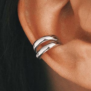 May include: A silver ear cuff with a double band design. The cuff is worn on the ear cartilage and has a smooth, polished finish. The image shows the ear cuff in detail, highlighting its sleek and modern aesthetic.