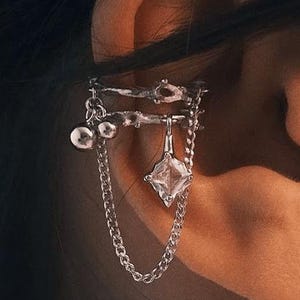 Pode incluir: Punho de orelha prateado com corrente e cristal pendurado. O punho de orelha envolve a orelha com pequenos enfeites redondos e um pendente de cristal maior, de corte quadrado. A joia foi concebida para ser usada numa orelha.