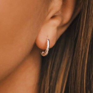 Puede incluir: Primer plano de un pendiente de aro plateado en la oreja de una persona. El pendiente tiene una superficie texturizada y es un diseño pequeño y curvado. La persona tiene el pelo largo y castaño y lleva una camiseta negra.