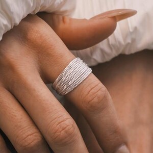 Puede incluir: Primer plano de dos manos adornadas con anillos de plata. Uno es una banda ancha con múltiples filas de pequeñas cuentas, el otro un delicado anillo de cadena con una cuenta colgante. Las manos tienen uñas largas y cuidadas y están sobre una chaqueta blanca.
