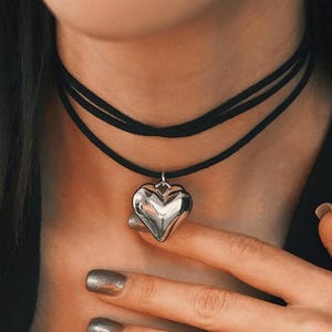 May include: A silver heart pendant necklace with a black choker. The heart pendant is a shiny, three-dimensional design. The necklace is worn around the neck, with a hand touching the necklace.