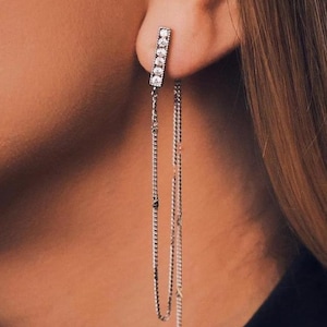 May include: A silver-toned earring featuring a rectangular bar stud set with five small clear stones. Two delicate silver chains hang from the base of the bar, extending downward to create a layered, dangling effect against the side of the head.
