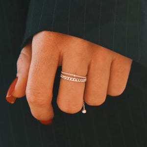 May include: A silver chain ring with a dangling charm is worn on a finger. The ring has two bands, one a delicate chain design and the other a plain band. The charm is a small, round bead. The background is a dark green suit.