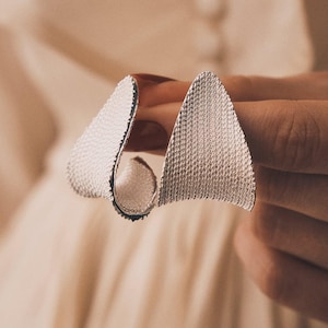 May include: A silver ring with a textured, geometric design. The ring features two large, triangular elements that curve around the finger. The surface of the ring has a detailed, dotted pattern. The ring is held up against a neutral background.