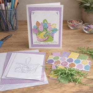May include: A handmade Easter card with a basket of colorful eggs, surrounded by craft supplies like scissors, glue, and decorative paper. The card is on a wooden surface, suggesting a crafting theme.