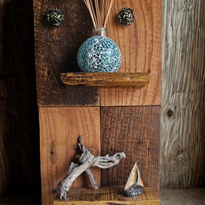 Puede incluir: Decoración de pared rústica de madera con un difusor de vidrio de mosaico y cañas, dos pomos decorativos, una pequeña repisa con madera flotante y un velero en miniatura. La madera tiene varios tonos marrones.