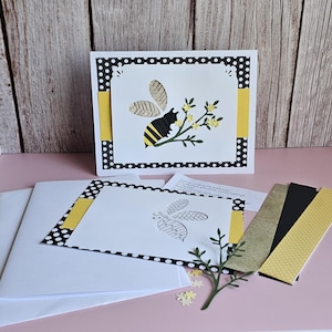Puede incluir: Una tarjeta de felicitación hecha a mano con un diseño de abeja. La tarjeta presenta un fondo blanco con un borde de lunares negros y detalles en amarillo. Una ilustración de abeja está sobre una rama con flores amarillas. Incluye sobres y papel decorativo.