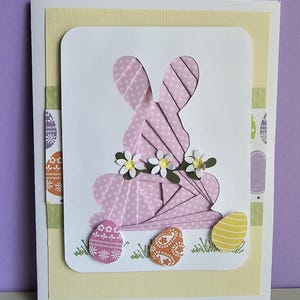 May include: Easter greeting card featuring a pink paper bunny silhouette with a polka dot pattern, adorned with small white flowers. The card includes decorative Easter eggs and green grass accents, set against a yellow and purple background.