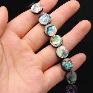 May include: A strand of circular abalone shell beads featuring a natural iridescent finish with swirling patterns of green, blue, purple, and pink hues. Each bead displays a unique, polished surface reflecting light with a pearlescent luster.