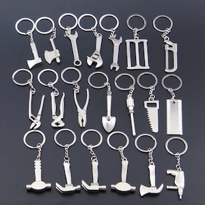 Peut inclure: Un ensemble de dix-huit porte-clés en métal argenté, chacun doté d'une breloque en forme d'outil miniature. Le lot comprend divers instruments tels que des marteaux, des clés, des pinces, des scies, une pelle, un tournevis, une hache et une perceuse, tous fixés à des anneaux fendus circulaires.