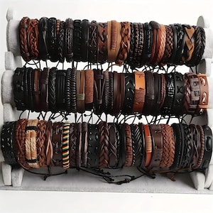 May include: A collection of numerous leather bracelets arranged on a white tiered display stand. The bracelets feature various designs including braided, woven, and smooth leather bands in shades of brown, black, and tan, with one featuring red, yellow, and green accents.