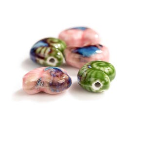 May include: A collection of five small, snail-shaped ceramic beads with a glossy finish. The beads feature a blend of pastel pink, vibrant green, and deep blue glaze colors, each with a central hole for threading, arranged in a cluster.