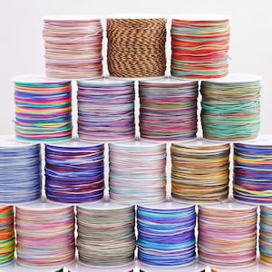 May include: A collection of spools of cord arranged in a pyramid formation. The spools feature various colors including solid light pink, black and brown twisted patterns, multi-colored stripes, and solid shades of blue, purple, green, and yellow.