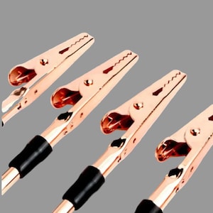 May include: Four rose gold metal alligator clips arranged diagonally. Each clip features a serrated jaw, a central pivot point, and a black rubberised sleeve covering the base of the handle. The metallic finish is reflective and smooth throughout.