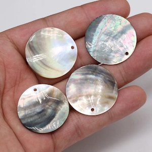 May include: Four circular mother-of-pearl shell pendants with iridescent surfaces. Each flat, round disc features a small pre-drilled hole near the edge for attachment. The natural shell material displays swirling gray, white, and pearlescent tones.