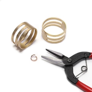 Puede incluir: Dos anillos de metal en tono dorado con un diseño de tres bandas y aberturas horizontales, acompañados por una pequeña anilla de salto color cobre y unos alicates con mangos negros y rojos para el montaje de joyería.