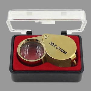 May include: A gold-colored jeweler's loupe with a 30X-21MM magnification lens, featuring a folding metal frame. The magnifying tool is housed inside a black rectangular plastic case with a clear hinged lid and a bright red velvet-lined interior.