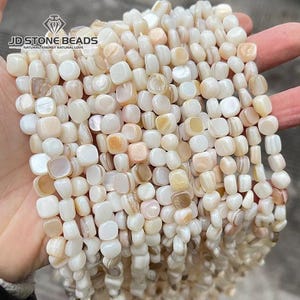 Op de afbeelding: Meerdere strengen vierkante, gebroken witte parelmoerkralen met een gladde, iriserende afwerking. De kralen vertonen subtiele variaties in crème- en lichtbruine tinten. De afbeelding bevat de tekst JD STONE BEADS NATURAL ENERGY NATURAL LOVE.