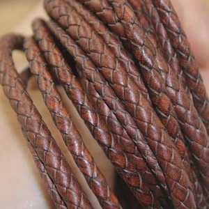 Puede incluir: Una longitud enrollada de cordón de cuero trenzado de color marrón oscuro. El material presenta un patrón de tejido apretado y consistente con una textura suave y mate, y variaciones de grano natural visibles en la superficie de los hilos.