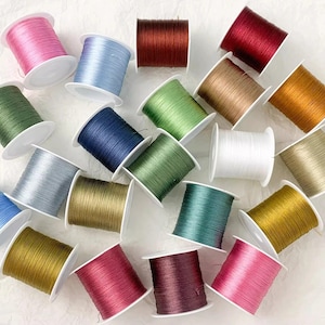 Pode incluir: Uma coleção de vinte e uma bobinas de linha em várias cores, incluindo rosa, azul claro, vermelho escuro, verde, azul marinho, branco, dourado, prateado e castanho. Cada bobina tem um núcleo de plástico branco com linha lisa e brilhante.