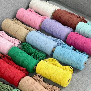 Pode incluir: Uma coleção de vários carretéis de corda de macramé em várias cores sólidas, incluindo branco, rosa, castanho, lavanda, azul claro, turquesa, magenta, verde, amarelo, vermelho e bege, dispostos num padrão de grelha sobre uma superfície cinzenta.