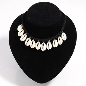 以下が含まれることがあります： 黒い編み込みコードのチョーカーネックレス。小さな銀色の金属リングで取り付けられたオフホワイトのタカラガイが並んでいます。貝殻は編み込みコードの前面に沿って一列に配置されています。