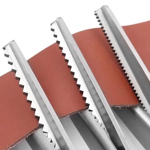 May include: Three silver-colored metal tools with serrated edges arranged in a parallel formation. Each tool features a long, straight spine and a row of sharp, triangular teeth along one side. A piece of smooth, reddish-brown leather is positioned underneath.