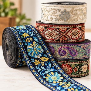 Könnte beinhalten: Ein Stapel aus fünf Rollen dekorativem Webband mit aufwendigen Stickereien. Die Muster umfassen blaue und gelbe Blumen auf Schwarz, rote und goldene geometrische Formen, violette Paisley-Muster sowie cremefarbene mit goldenen Blumen.